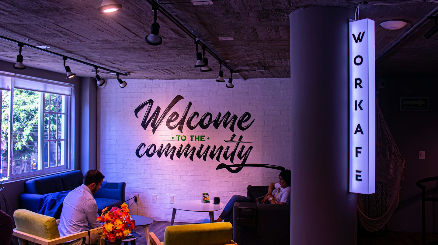 Two people relax in a co-working cafe with a "Welcome TO THE community" mural.